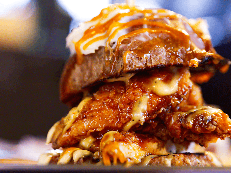 Close-up of a tall fried chicken burger with glossy bun and dripping sauce