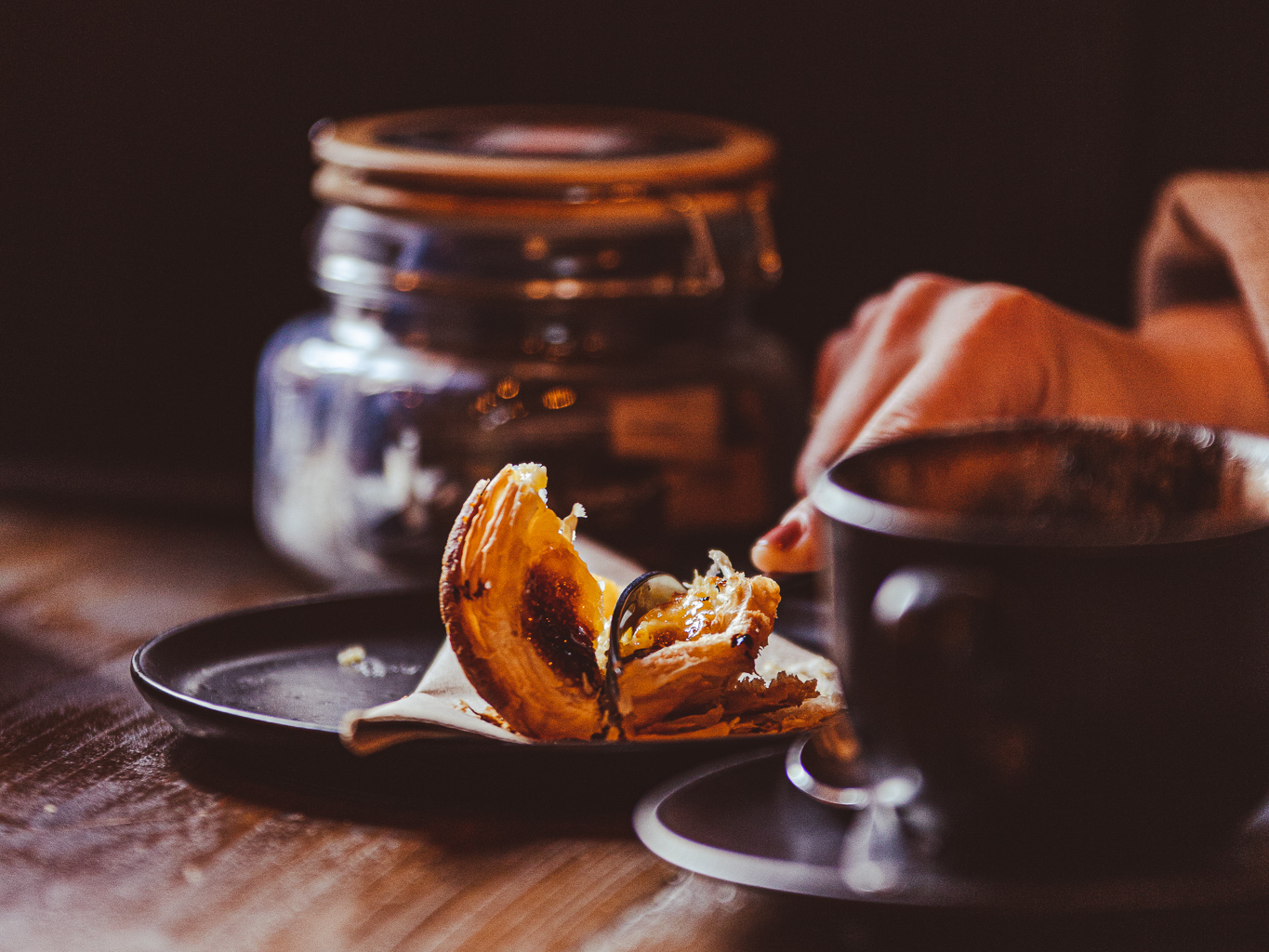 Close-up of a pastel de nata on a dark plate with a coffee in warm café lighting